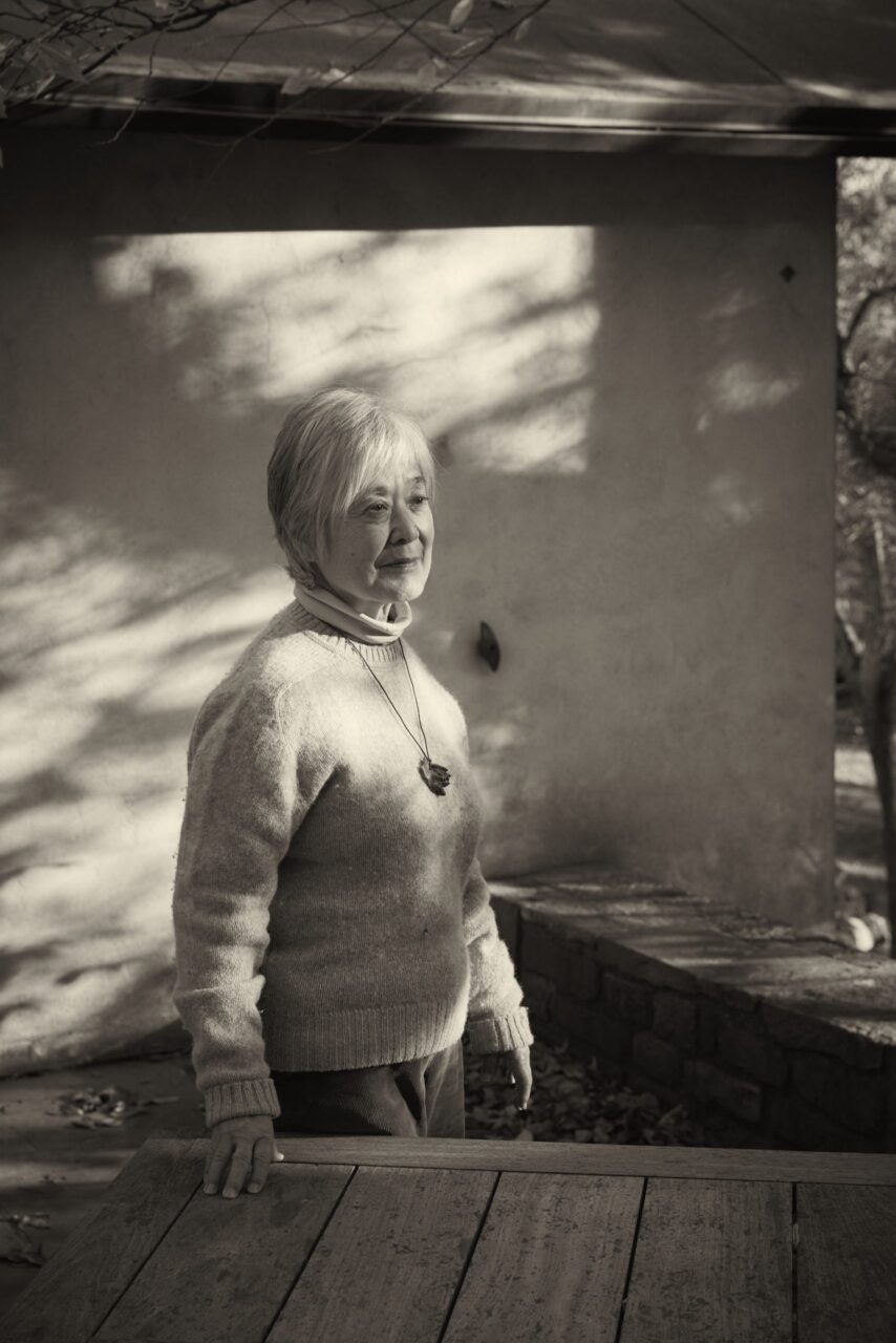 Monochromatic image of Mira Nakashima standing in dappled light in front of the Nakashima Chair Studio. Photo credit Martien Mulder.